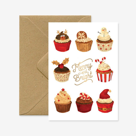 A Christmas-themed greeting card with various illustrated cupcakes and the festive greetings 'Merry & Bright' written on it, accompanied by a brown envelope.