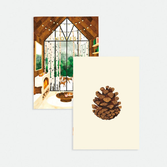 A pocket notebook with a gold foil cover featuring an illustration of a pine forest and a cabin. The notebook is blank and has a cream-colored spine.