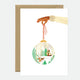 A Christmas greeting card featuring a hand holding a round ornament with a forest and snowscape scene inside, accompanied by a brown envelope.