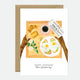A birthday greeting card with a illustration of a breakfast plate and a cup of coffee, with gold foil details and the text 'Happy Birthday' on the front.