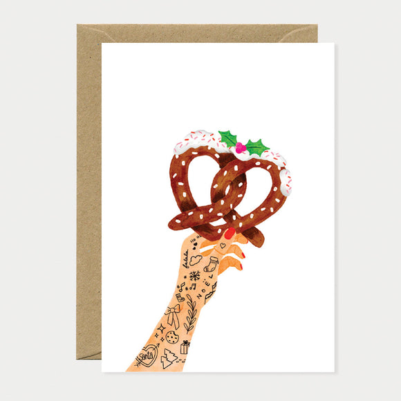 Greeting card with a tattooed hand holding two sugar-topped pretzels decorated with holly leaves for a festive Christmas design.