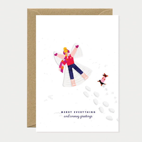 Greeting card showing a child making a snow angel while a dog plays nearby, creating a joyful festive winter scene.