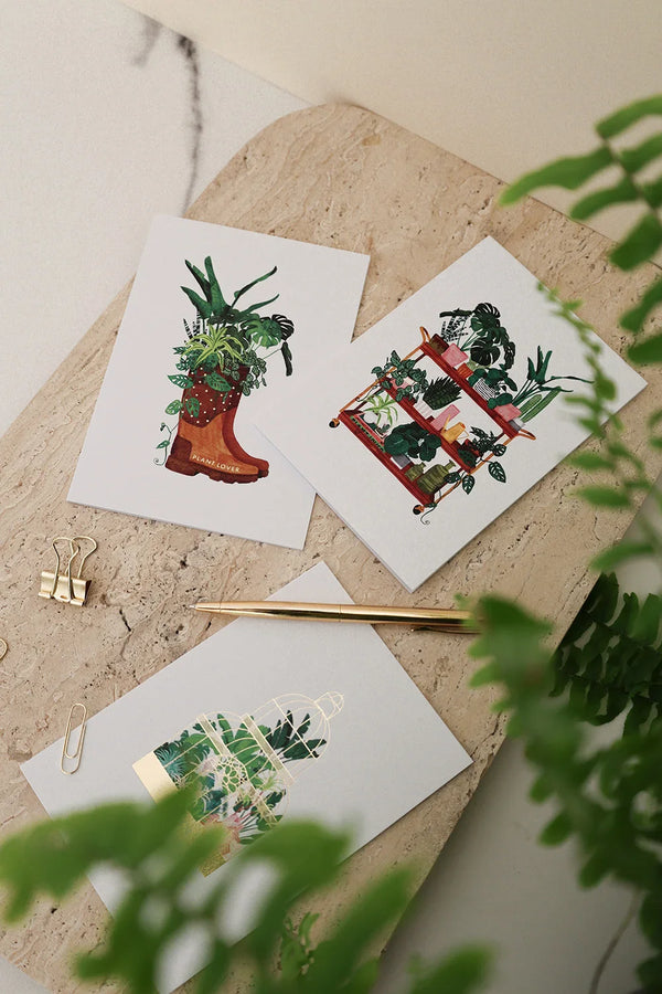 Set of illustrated botanical greeting cards featuring plants in a boot, greenhouse, and shelf, styled with gold pen and clips.
