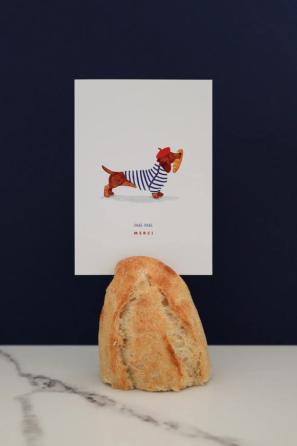 All The Ways To Say Oui Oui Merci card – dachshund in striped shirt holding baguette, styled with bread and marble backdrop