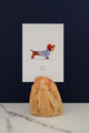 All The Ways To Say Oui Oui Merci card – dachshund in striped shirt holding baguette, styled with bread and marble backdrop