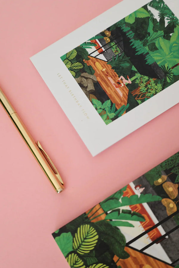 Birthday card featuring a tropical yoga scene with a woman practicing, styled on a pink background with a gold pen.