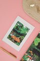 Tropical birthday card with jungle illustration and woman doing yoga, placed on a pink background with gold pen and binder clip.