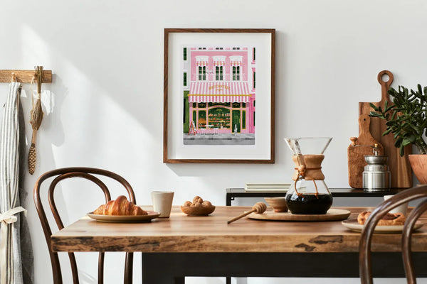 Pink Bakery art print framed in a bright kitchen with wooden table, croissants, and coffee