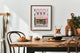 Pink Bakery art print framed in a bright kitchen with wooden table, croissants, and coffee