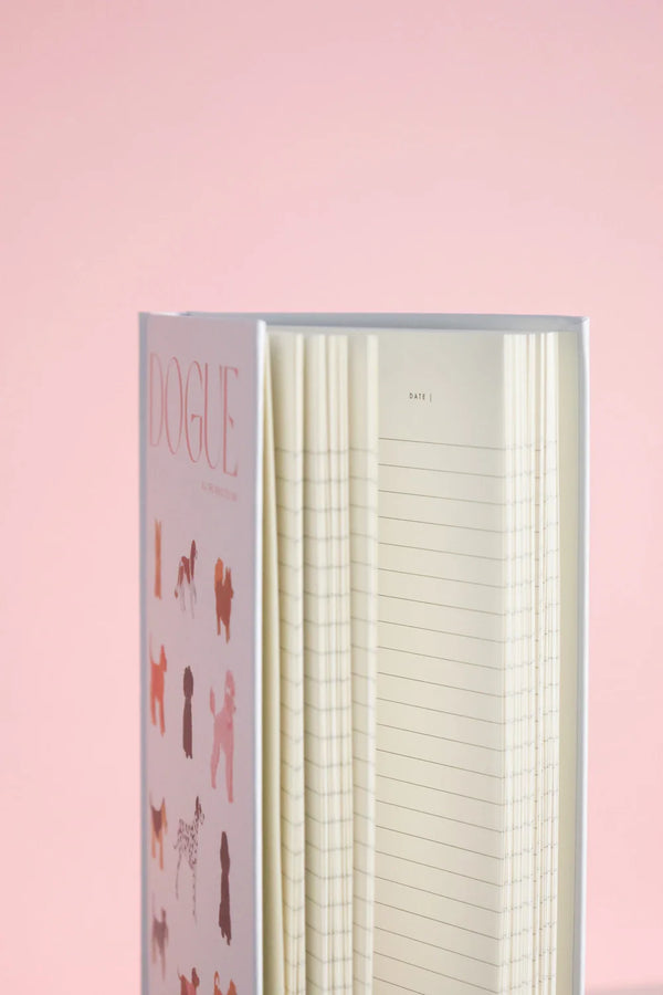 Side view of Dogue notebook spine with pink title and lined interior pages against soft pink background.