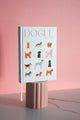 Dogue illustrated dog notebook displayed upright on pink pedestal with pink background, modern stationery design.