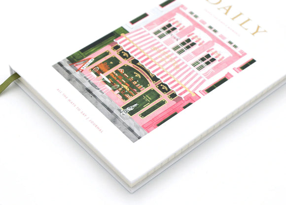 Daily hardcover notebook with bakery illustration laying flat, showing gold foil title and green ribbon bookmark.