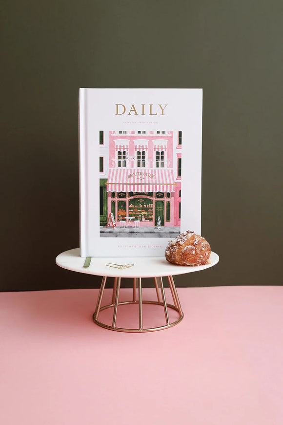 Close-up of the Daily Journal “Bakery” by All the Ways to Say, featuring a pink patisserie illustration with gold foil details, displayed on a marble stand with a choux pastry.