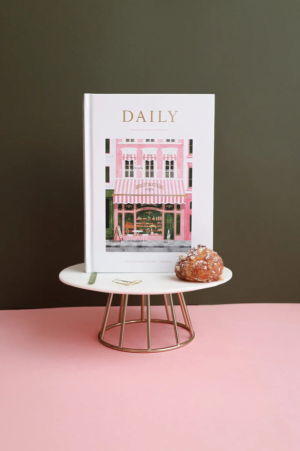 Close-up of the Daily Journal “Bakery” by All the Ways to Say, featuring a pink patisserie illustration with gold foil details, displayed on a marble stand with a choux pastry.