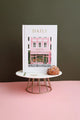 Close-up of the Daily Journal “Bakery” by All the Ways to Say, featuring a pink patisserie illustration with gold foil details, displayed on a marble stand with a choux pastry.