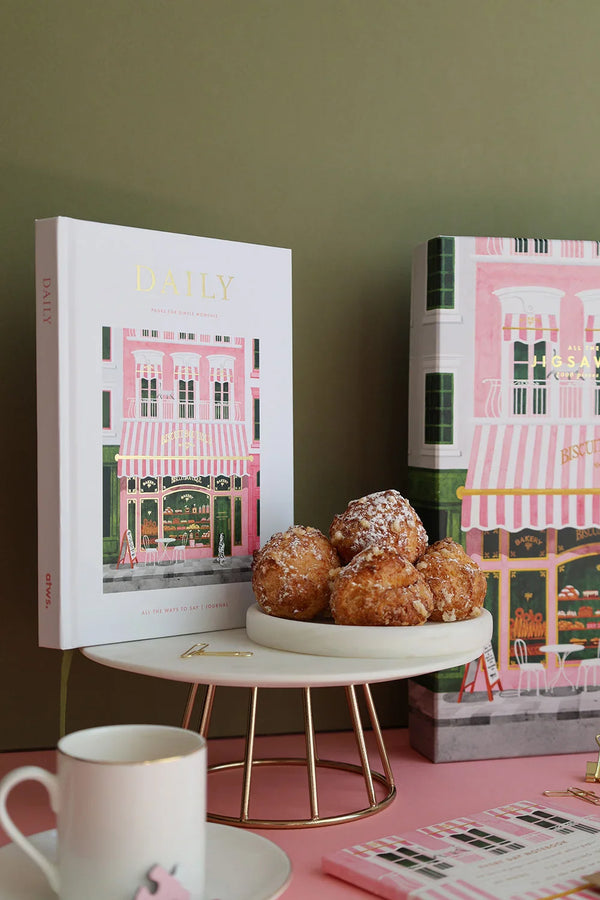 White coffee cup with espresso placed on the pink Bakery Daily Journal by All the Ways to Say, lit by natural sunlight.