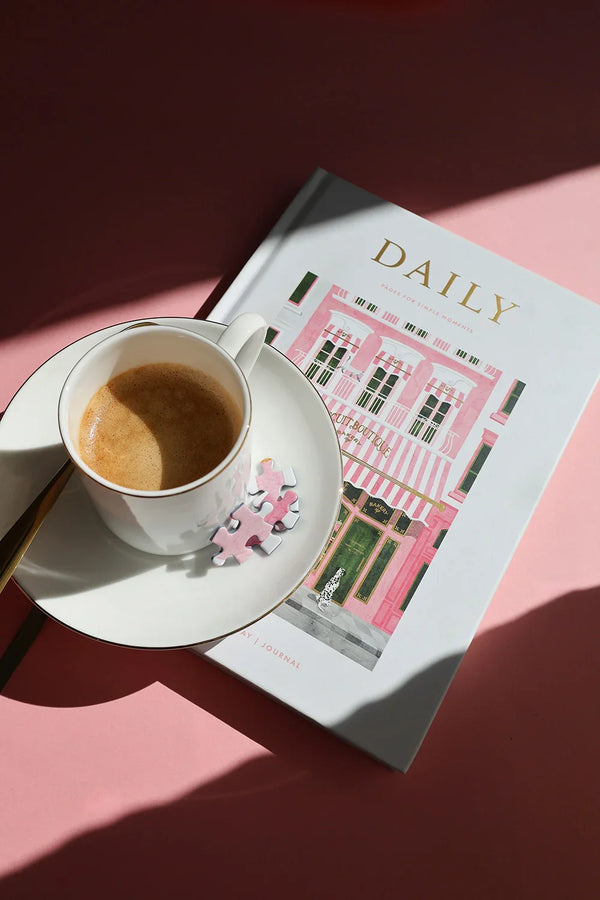 Cup of espresso with puzzle pieces resting on the Bakery Daily Journal, photographed in sunlight on a pink background.