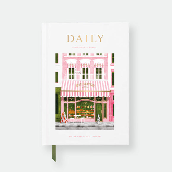 Hardcover notebook A5 with white cover, Paris bakery storefront illustration in pink and green, gold foil “Daily” title and green ribbon bookmark.
