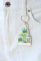 Green House keychain by All The Ways To Say hanging on white denim pocket – botanical enamel design