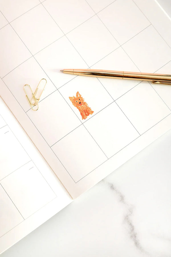 Interior of the Dogue monthly planner with a cute orange dog illustration and gold pen on a white marble surface.