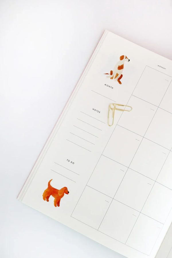 Page spread of the Dogue monthly planner with watercolor dog drawings, gold paper clips, and clean grid layout.