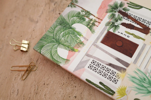 Close-up of an illustrated notebook cover with palm trees and modern house design, styled on a marble surface with gold paper clips.