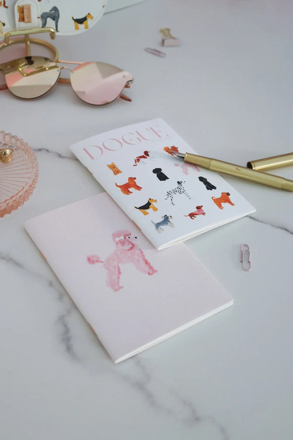 Dogue notebook with illustrated dog breeds on white cover, styled with gold pen and pink sunglasses for a chic desk setup.