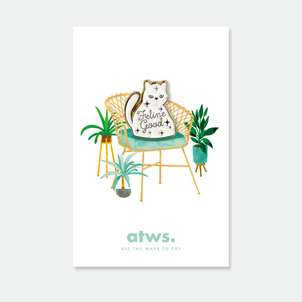 Feline Good enamel pin by All the Ways to Say featuring white cat with “Feline Good” text, gold-plated details, displayed on cozy chair with green plants background.