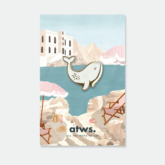 White Whale enamel pin by All the Ways to Say featuring pearly white whale with gold-plated details on illustrated seaside background with umbrellas.
