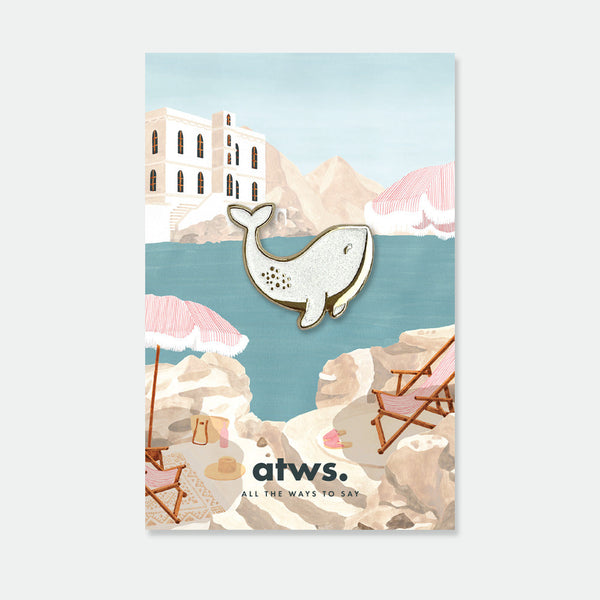 White Whale enamel pin by All the Ways to Say featuring pearly white whale with gold-plated details on illustrated seaside background with umbrellas.