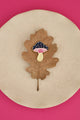 Crazy Mushroom enamel pin by All The Ways To Say on brown oak leaf over beige plate – pink background