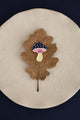 Crazy Mushroom enamel pin by All The Ways To Say on oak leaf over beige plate – navy blue background, minimal fall style