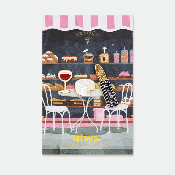 French Kit enamel pins by All the Ways to Say featuring wine glass, cheese wheel and baguette with gold-plated details on Parisian café background.