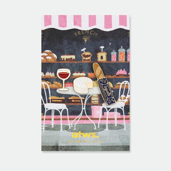 French Kit enamel pins by All the Ways to Say featuring wine glass, cheese wheel and baguette with gold-plated details on Parisian café background.