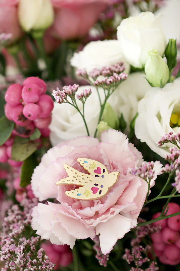 Lovebird enamel pin by All The Ways To Say placed inside pink carnation surrounded by white and pink flowers