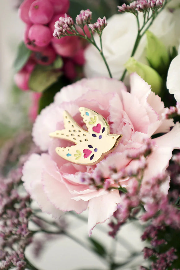 Close-up of Lovebird enamel pin by All The Ways To Say – gold bird with colorful hearts resting on pink carnation