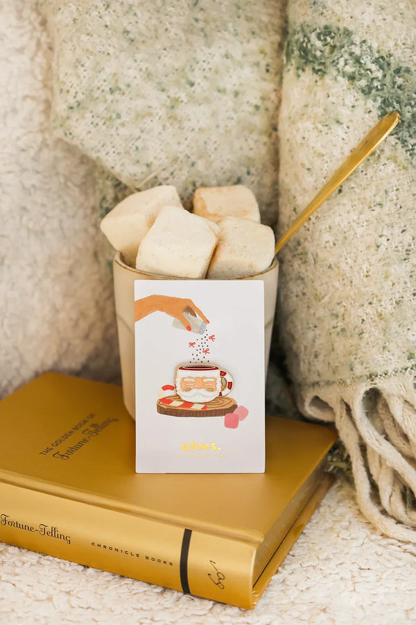 Santa’s Cup enamel pin by All The Ways To Say on illustrated card inside cozy winter setup with marshmallows and blanket
