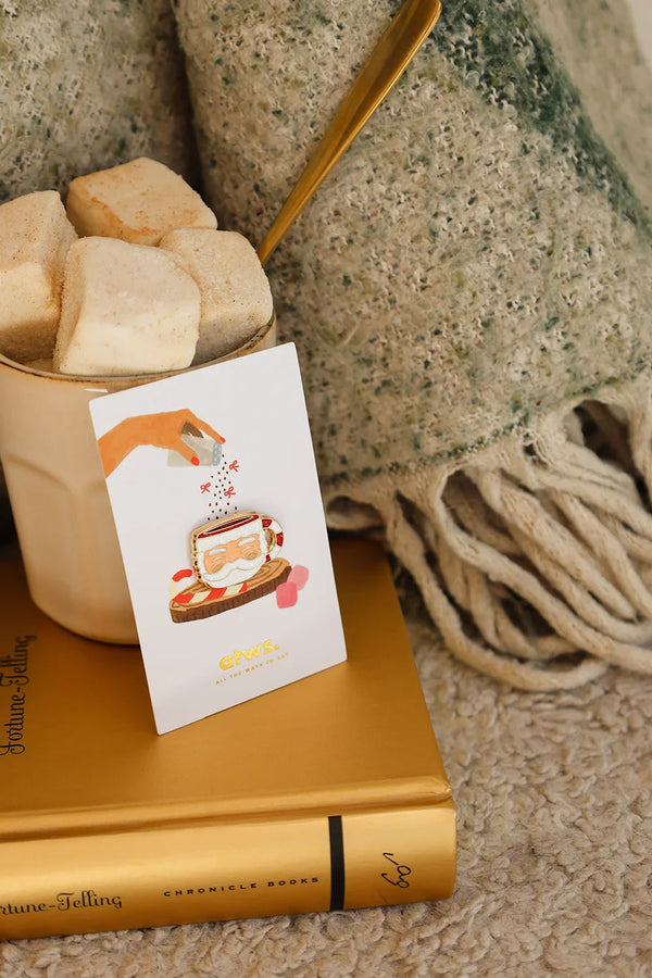 Santa’s Cup enamel pin by All The Ways To Say on beige mug filled with marshmallows – festive and warm Christmas scene