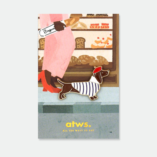 Little Frenchie enamel pin by All the Ways to Say featuring dachshund wearing red beret and striped shirt, gold-plated details on Paris bakery background.