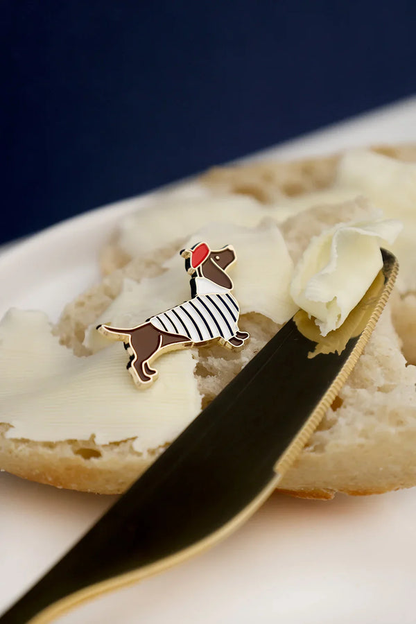 Close-up of Little Frenchie enamel pin by All The Ways To Say – dachshund dog in striped shirt and red beret on bread background