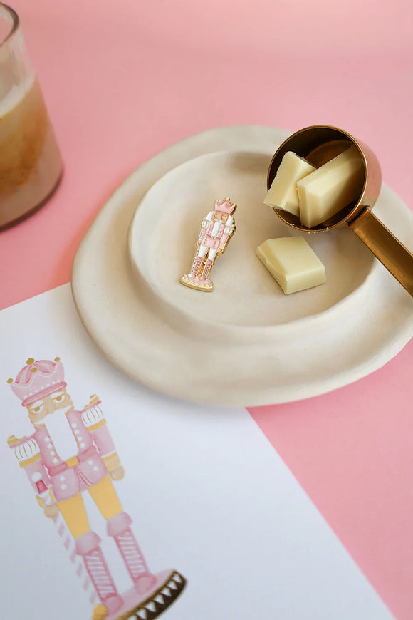 Pink Nutcracker enamel pin by All The Ways To Say on beige ceramic plate with white chocolate pieces – festive pastel setup