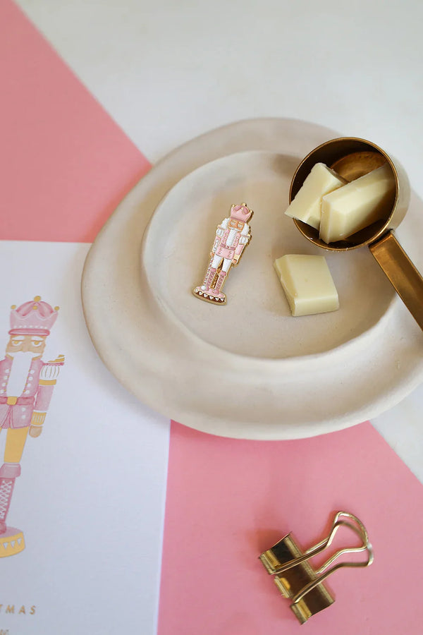 Pink Nutcracker enamel pin by All The Ways To Say on pink and white background – cozy Christmas mood with chocolate and gold details