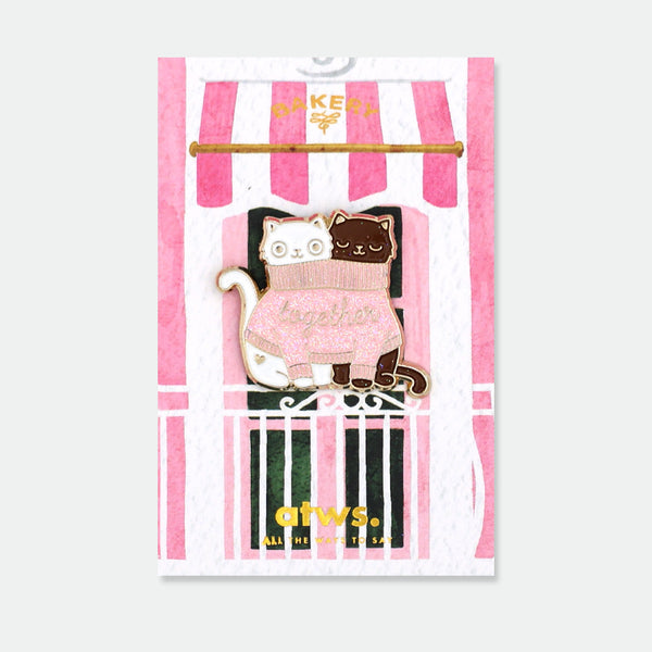 Together Cats enamel pin by All the Ways to Say featuring two cats sharing a pink glittery sweater with “together” text, gold-plated details on bakery background.
