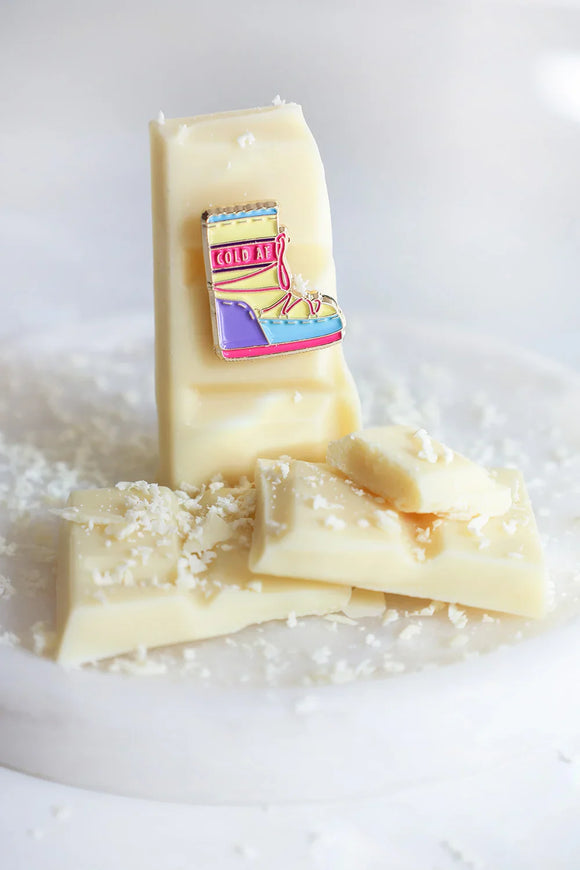 Close-up of Cold AF enamel pin by All The Ways To Say – pastel snow boot on creamy chocolate bar surface
