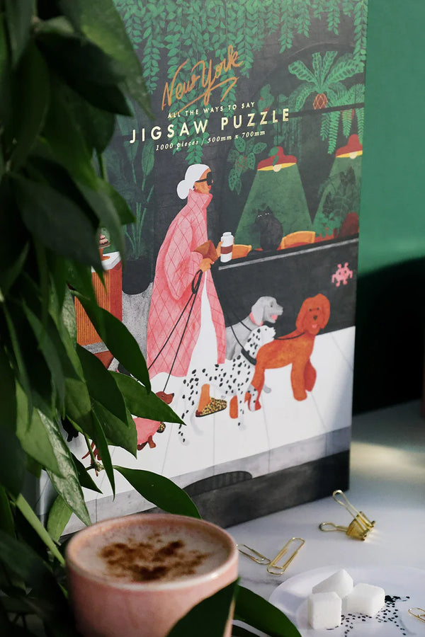 Lifestyle photo of The Dog Sitter puzzle – box, coffee cup, and scattered jigsaw pieces on white marble.