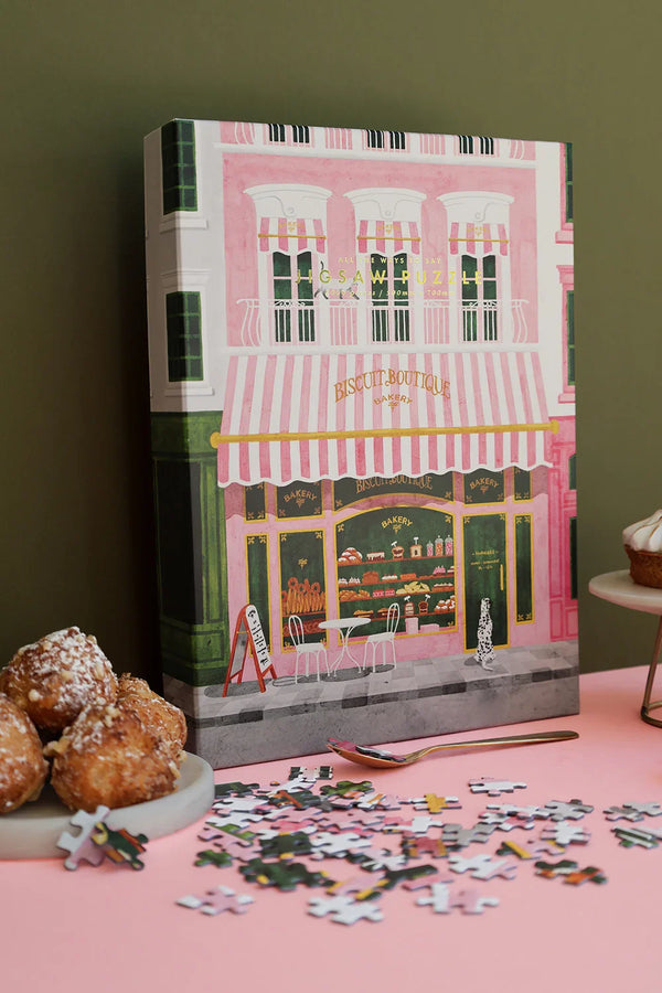Puzzle pieces scattered on a pink surface beside the Pink Bakery puzzle box — elegant café illustration in soft pink and green.