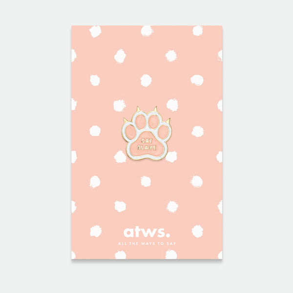 A gold plated enamel pin in the shape of a cat paw, displayed on a card with a pink polka dot background.