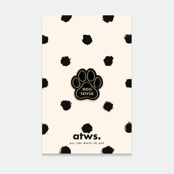 A packaged enamel pin with a dog paw design and the text 'DOG LOVER' on it, predominantly gold and black in color, presented on a card with black polka dots and the brand 'atws' at the bottom.