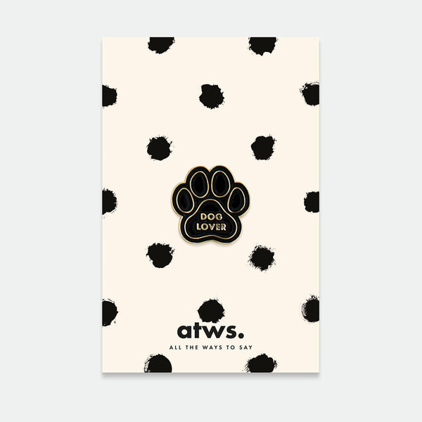 A packaged enamel pin with a dog paw design and the text 'DOG LOVER' on it, predominantly gold and black in color, presented on a card with black polka dots and the brand 'atws' at the bottom.