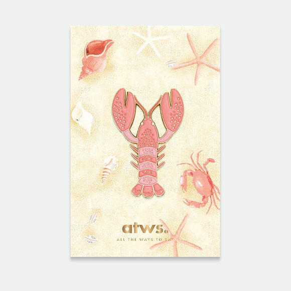 A card with a gold-plated enamel pin depicting a lobster, surrounded by starfish and shells on a beige background.